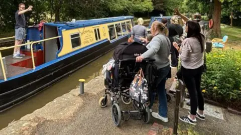 A blue and yellow boat on a canal with a number of people, one of them in a wheelchair, waving from the towpath