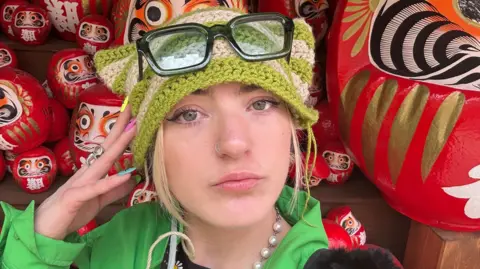 Izabel Rose, a woman with blonde hair and hazel green eyes looks into the camera - she wears a knitted green and beige stripes hat with glasses perched on top and there are Japanese figures in the background