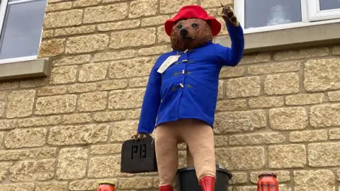 A scarecrow of Paddington Bear. He is wearing a red hat and blue coat and carrying a briefcase. There are jars of marmalade on the ground next to him. The scarecrow is positioned with an arm in the air in front of a brick building.
