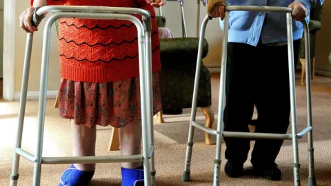 PA Media A generic shot of two elderly people with walking frames - their heads are not in shot. One is wearing a red and black striped jumper and a red skirt with a pattern and blue slippers. The other is wearing a blue cardigan and black trousers and slippers. They are side by side in a room with a light brown carpet. 