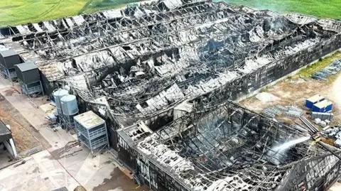 Cheshire Fire and Rescue Service An aerial image, taken from a drone, of a large farm building destroyed by a fire. The roof has caved in, leaving the building's skeleton exposed, and smoke is emanating from the site.