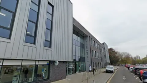 Google Maps A modern building composed of three stories with large windows and grey cladding. There is a vertical sign that reads "Quayside House" and the building overlooks a large car park. It is a grey and cloudy day.