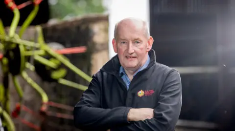 Glenn Cuddy is bald and has his arms folded. He's wearing a black jacket that says Ulster Farmers' Union in red lettering on the left breast. Under the jacket he is wearing a blue checked shirt.