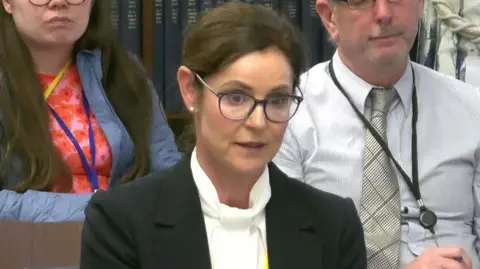 BBC Ciara Fitzpatrick giving evidence to the committee. She has brown hair that's tied back, and is wearing glasses, a black jacket with a white top, and silver earrings.