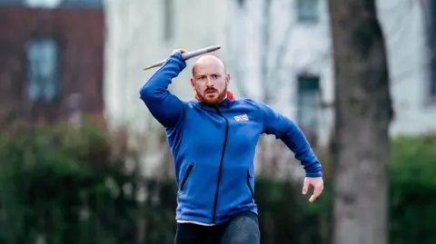 Dan Pembroke, in his run up, holding a javelin wearing a blue GB hoodie