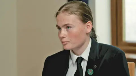 Bethany speaks to the BBC while wearing her school uniform, which includes a dark blazer with a green pin-on badge.