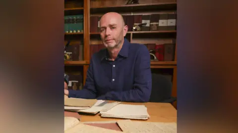 Trinity College Dublin Dr Gurrin is sitting at a desk with papers around him. He is looking at the camera and is bald. He has a navy shirt on and one arm is resting on the table.