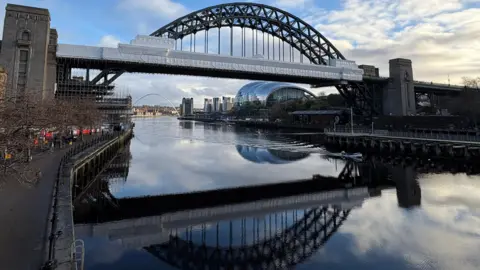 Tyne Bridge - the arch above the river is visible with scaffolding covering the roadway. There are trees on the river bank and buildings including in the distance. There is another bridge in the distance.