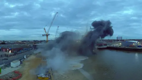 Sky Revolutions Smoke over the River Yare in the aftermath of a bomb blast, with a yellow crane visible behind it 