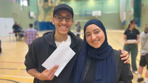 BBC Kareem has his arm around Jana. He is holding his GCSE results envelope. They are both smiling and are in a school sports hall