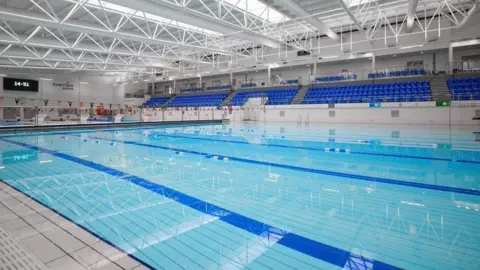 A wide angle shot of a swimming pool, with its blue flooring, and in the background is the blue seating in the gallery. There is a white-painted roof above.
