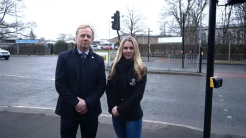 The King's School George Hartley and Claire Taylor stand at the side of the road by the crossing outside the school. Mr Hartley is wearing a dark jacket, blue shirt and green tie and dark coloured trousers. Mrs Taylor wears a dark coloured blazer with jeans and a black scarf.. 