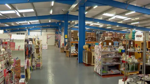 BBC The Acorn shop is in a large modern building. There are many shelving units holding vases, clothes, books and furniture.