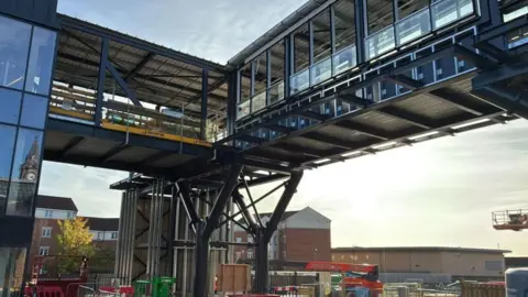 Network Rail A modern covered footbridge is held up by a big pillar as it switches direction by ninety degrees and enters a modern building. Behind are some new housing developments. 