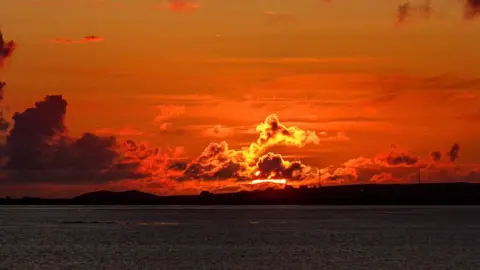 Dória de Souza Etchells A bright red and orange sunset glows from behind dark clouds over the water.