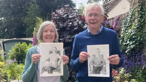 A woman and a man stand in a colourful garden while smiling and holding A4-sized black-and-white portraits of themselves. The woman has grey hair in a bob and is wearing a green cardigan over a white and burgundy striped shirt. The man has short white hear and is wearing glasses and a blue jumper over a light blue shirt. The garden has green and red shrubs along with colourful climbing plants against a dark brick wall.