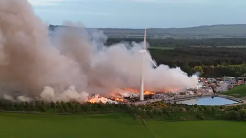A landfill site next to some water and a windmill is on fire with smoke rising from the scene.