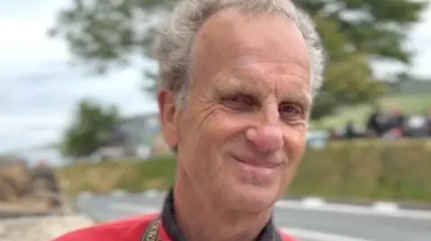 John Watterson John Watterson, who has grey wavy hair and is wearing a red jacket, standing at the side of the TT course.