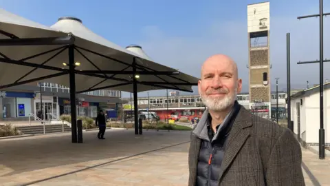 A man in a Tweed jacket standing next to a canopy and open paved space with a concrete clock tower in the background 