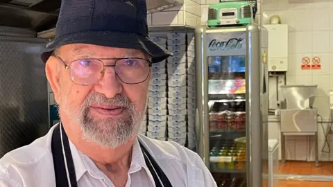 John Panayis in a white shirt, a black apron and black hat stands in front of frying equipment.