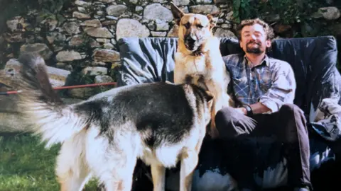 Mick Murray Bob sits outside behind a stone wall, sharing a chair with two Alsatian dogs. He smiles at the camera.