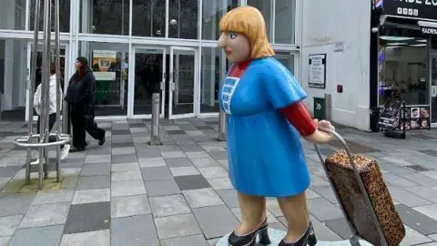 Sculpture of a woman wearing a blue dress pulling a trolley on a pavement
