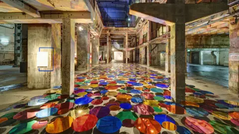 Klaus Bossemeyer The interior of Cabot Circus multi-storey car park. It is a large industrial building with concrete walls, floors and columns. It has been filled with colourful acrylic disc on the floor, and overhead lighting is casting beams of light over the walls.