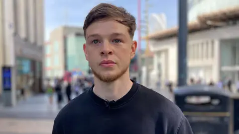 Rudy O’Halloran is looking out of shot, in the centre of Birmingham. He has brown hair and a small beard and he is wearing a blue sweatshirt