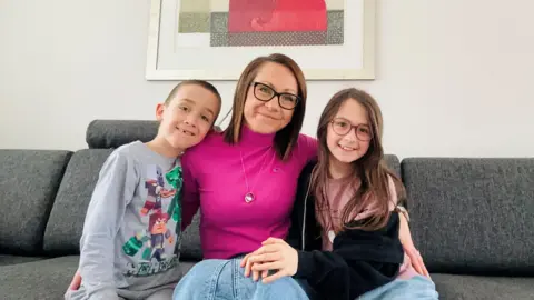 Jojo Clark A woman with short brown hair and a bright pink jumper sits on a grey sofa with a framed, abstract painting on the wall behind her. Her young son sits to the left of her wearing a grey Minecraft t-shirt and her daughter is on the right wearing a light pink top and an unzipped, dark-coloured hoodie.