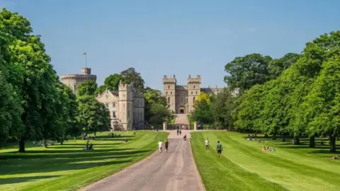 Windsor Castle is pictured at the end of a long, straight pedestrian thoroughfare called the Long Walk. Neatly trimmed grass is on either side, which is flanked by trees. 