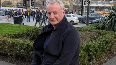 A man in his sixties sitting on a wall, behind which a small hedge plant sits. In the background is Sheffield Station.