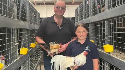 Alannah (right) stands with her dad Neal (left).Neal is wearing a black polo shirt and jeans and glasses. Alannah is wearing a navy polo shirt with a white and green logo. Neal is holding a small brown and black chicken and Alannah is holding a larger white chicken with long tail feathers. They are both smiling at the camera. They are standing inside an indoor chicken coop with multiple enclosures. There are metal grated gages with bright yellow feed boxes on the front.