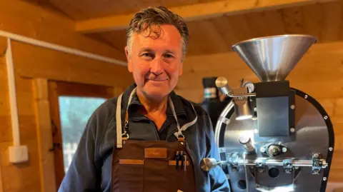 BBC A man with grey hair, wearing a brown apron and blue jumper and smiling at the camera. Standing next to a silver coffee roasting machine. He is in a wooden out building