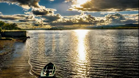 Rays of sunshine hit the water despite mostly dark clouds in the sky. A small rowing boat is sitting in the bay. 