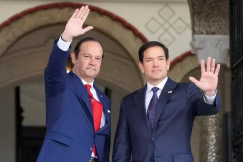 Reuters US Secretary of State Marco Rubio waves with Panamanian foreign minister Javier Martinez-Acha after arriving at the presidential palace in Panama City. Both men are wearing blue suits.