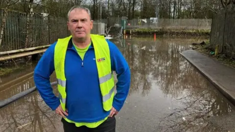 Carl Belford of Belford Brothers Skip Hire, wearing a blue jumper and a yellow high visibility jacket. He is standing at the edge of a flooded road. There is brown flood water and bits of debris behind him.