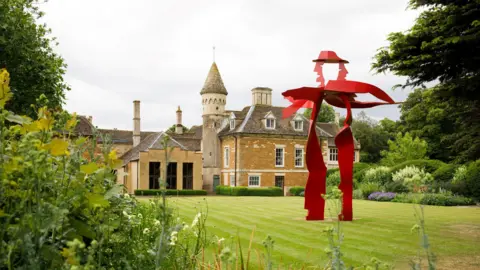 Kieran Vyas A photo of an old stately home in Leicestershire with honey coloured stone with a striking red modern sculpture in the foreground. In the background, lush mature gardens are filled with white, purple and red flowers.