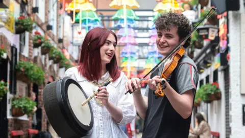 A woman and man standing in Belfast's Cathedral Quarter. The woman, with dark red hair, is holding a bodhrán. The man, with short, curly brown hair, is holding a fiddle. 