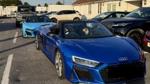 A dark blue Audi supercar, with a light blue supercar and a large grey 4x4 parked on the forecourt of a small building. 