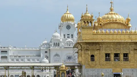 A view of the Golden Temple complex in Amritsar, Punjab. The gold-covered Harmandir Sahib dominates the foreground, while white marble buildings with domes and a clock tower rise behind it under a clear blue sky. The temple is the holiest shrine in Sikhism and is visited by millions of pilgrims each year.