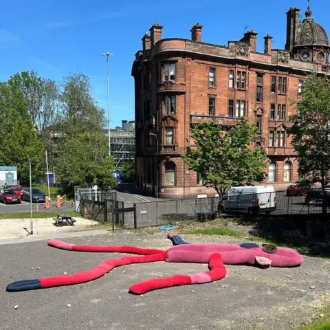 Glasgow School of Art A sculpture of a giant sock puppet lying in a car park. It has a pink body, one blue and one red arm and two red legs. It has two giant button shapes for eyes.