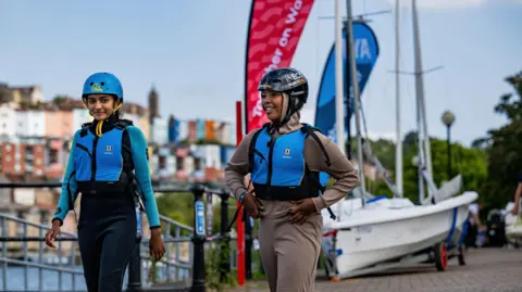 Royal Yachting Association/Martin Allen Photography Two girls walking along the the harbour with a small sailing boat behind them. They're wearing blue lifejackets and helmets