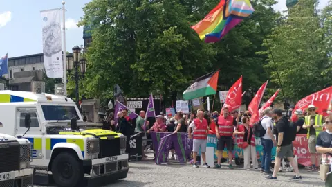 Police vehicles and protesters lined up outside Belfast City Hall during an anti-racism protest.  Two police Land Rovers are visible in the foreground.  The protesters are waving flags including red Nipsa flags. 