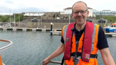 Tom Willis, a middle-aged man with glasses, stands on the deck of the boat smiling at the camera wearing a lifejacket and hi-vis.