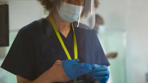 Getty Images Medical professional wearing dark blue scrubs with a yellow lanyard. She is wearing a face covering including visor and in the process of putting on protective gloves