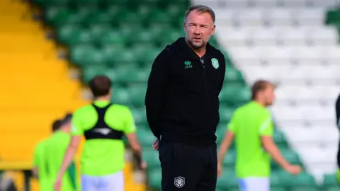 Marcus Stewart is standing on a pitch in a black outfit. Behind him, players are training in high-vis, green shirts. The bleachers are blurry in the background.