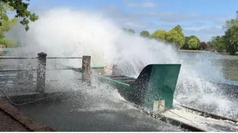 A green boat makes a large splash next to a wooden jetty - the boat has two passengers but could hold eight.