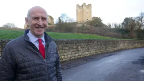 John Healey A man with a bald head is wearing an black padded jacket, a white shirt and red tie. He is smiling and standing in a street with a castle turret in the background