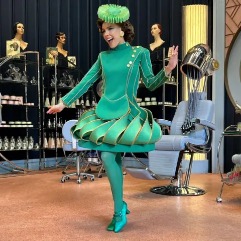 Lily Field Lily Field wearing an Emerald City costume in the film Wicked. She is striking a pose in a scene in a hairdressing salon.