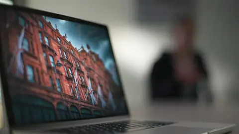 Sarah is seen heavily blurred to protect her anonymity. She is sitting in an office with a laptop in the foreground and an image of the exterior of Harrods on it